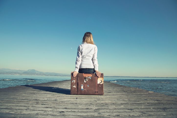 Rear view of person with long blonde hair sitting on suitcase at the end of a dock