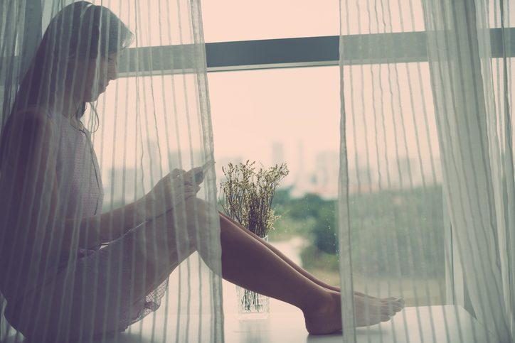 Young woman sitting on windowsill behind curtains and listening to the music