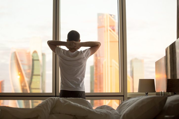 Man wearing t-shirt and sweats looks out window of high-rise building, hands clasped behind head