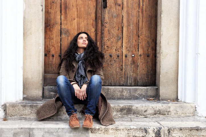 Person with long hair sits on stairs outside old wooden door, looking up with disappointed expression