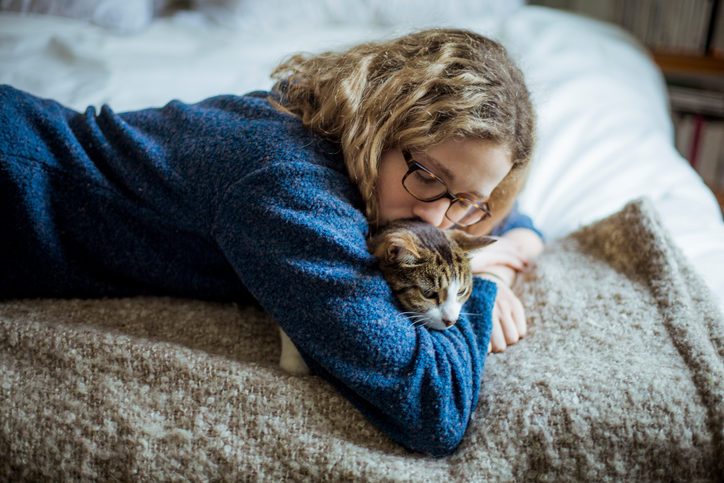 Person in blue sweater with long wavy blonde hair and glasses lies on bed with somber expression holding cat and cuddling