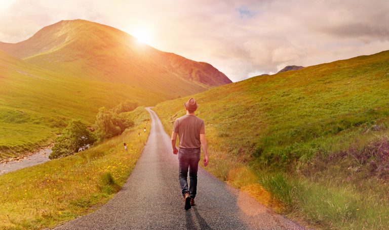 Rear view of person in casual clothes and hat walking alone on a narrow country road through a green valley surrounded by mountains, heading toward the rising sun. Warm sunlight with lens flare. Taking decisions and choosing a direction for new beginnings, following the way forward, not looking back.