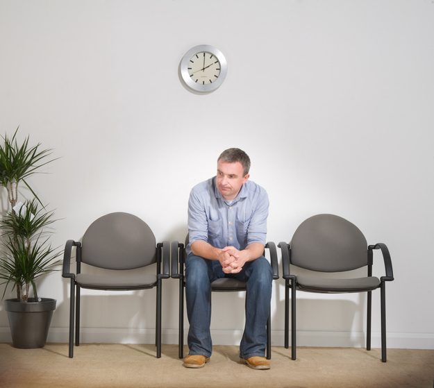 Bored-looking person sits alone in waiting room, leaning forward, hands folded