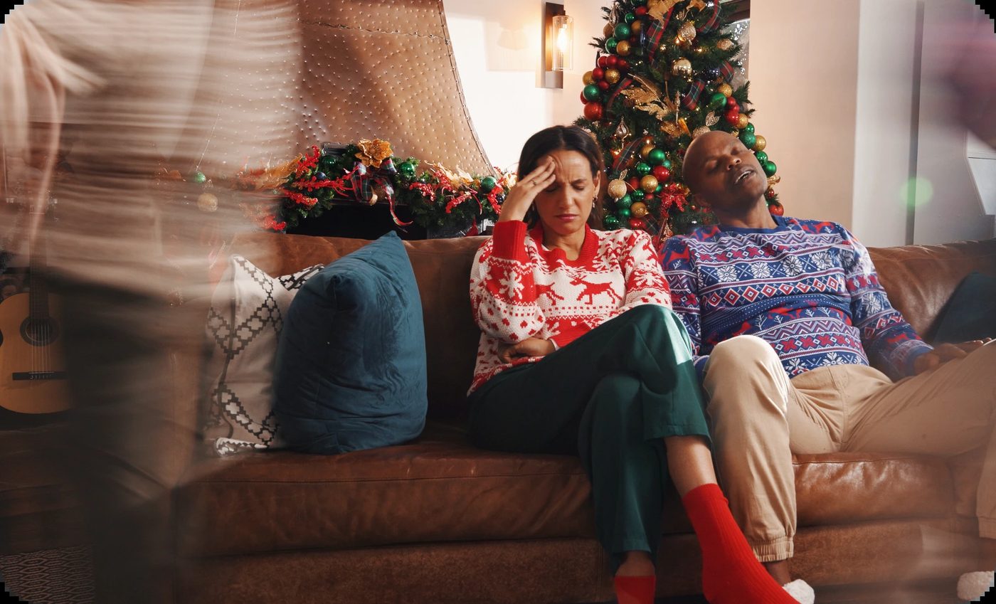 A stressed woman with a hand on her forehead and a sleeping man on a couch, surrounded by festive holiday decor, illustrating understanding winter depression.