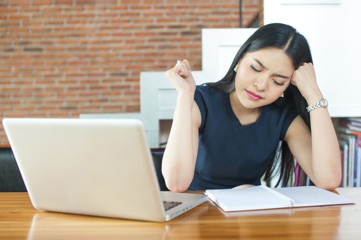 Young professional sits in front of laptop with open notebook, head on hand, looking bothered