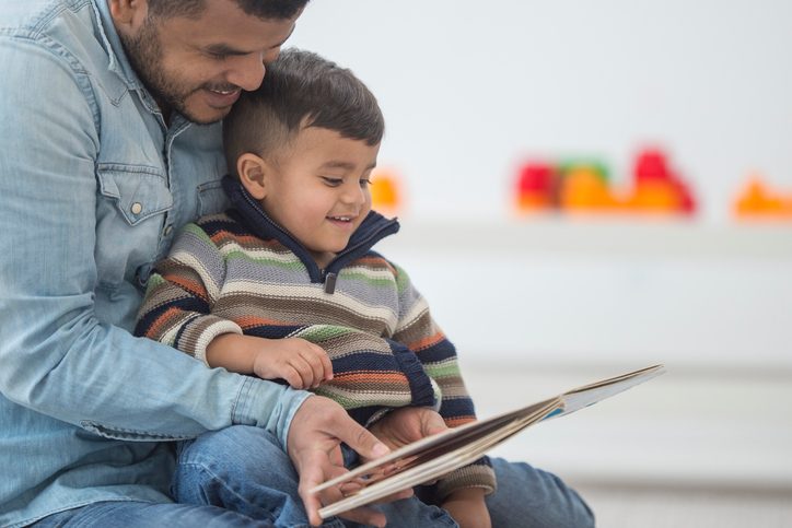 Young child sits in lap of parent with short hair who reads large picture book. Both parent and child are smiling