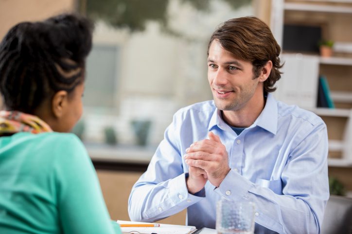 Attentive therapist listens to young adult seated at table with a smile and hands clasped