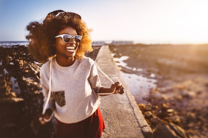 Photo of young adult with brilliant smile laughing and looking off to one side, holding backpack straps out, standing on walking trail