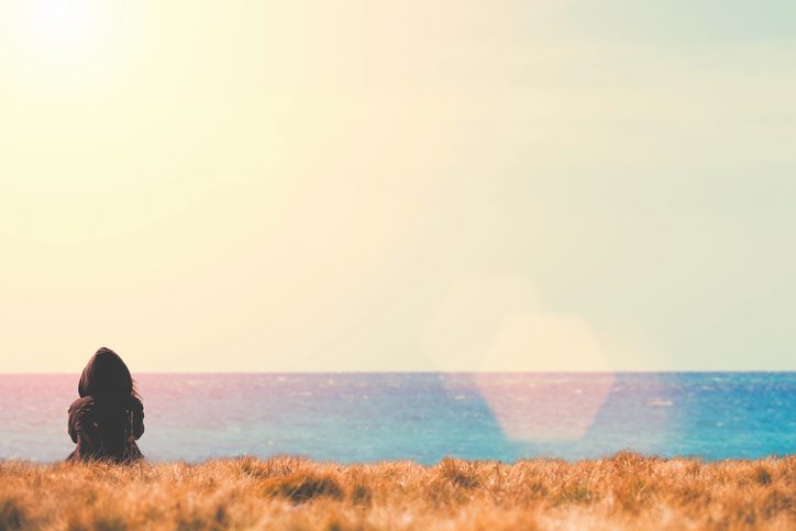 Rear view of person sitting on grassy beach area looking out toward the water and horizon