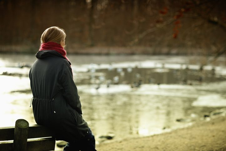 Person wearing red scarf and black coat sits on back of bench and looks out at lake