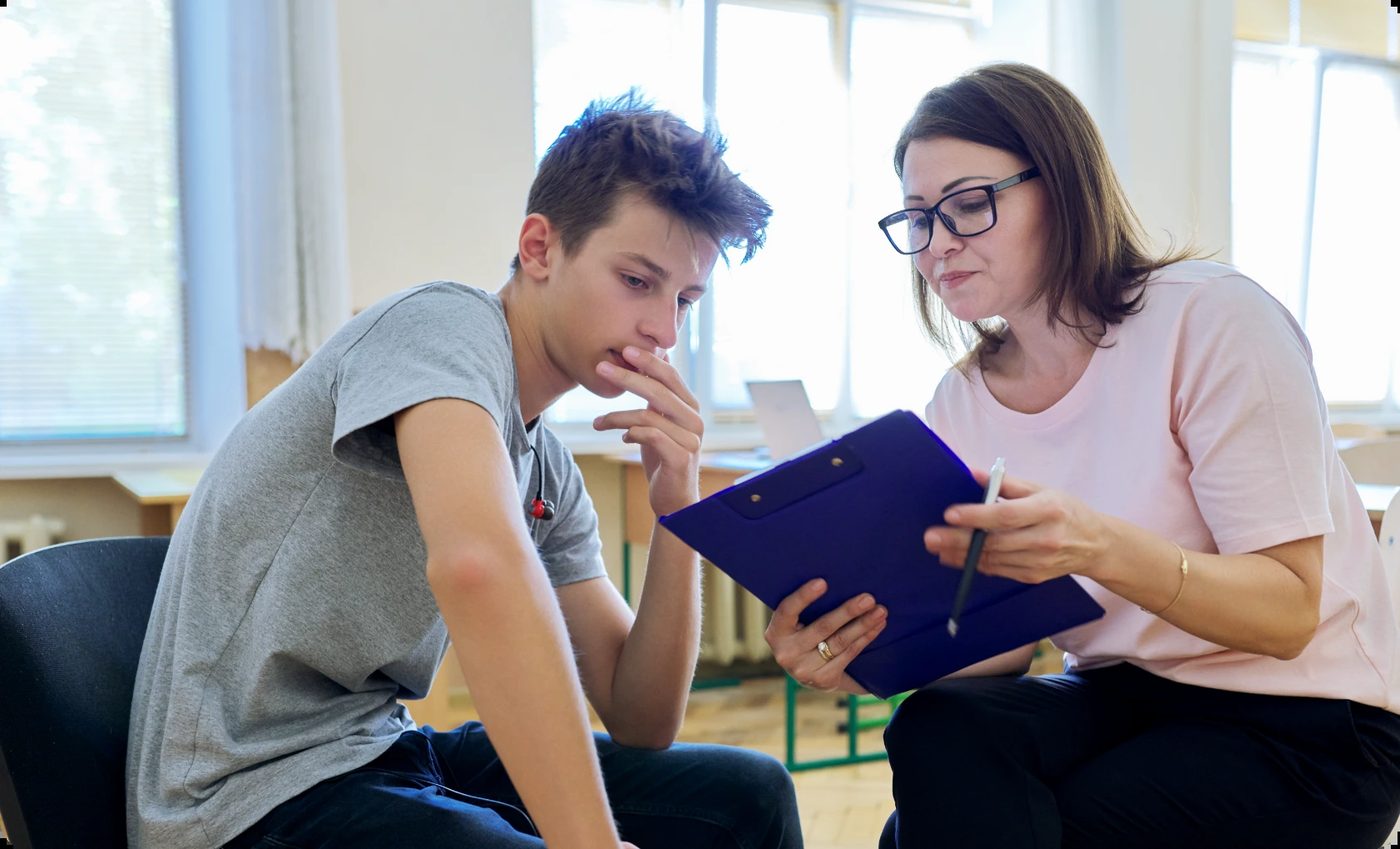 Female therapist with glasses and a young male client sit together reviewing a clipboard during a types of therapy session in a bright room