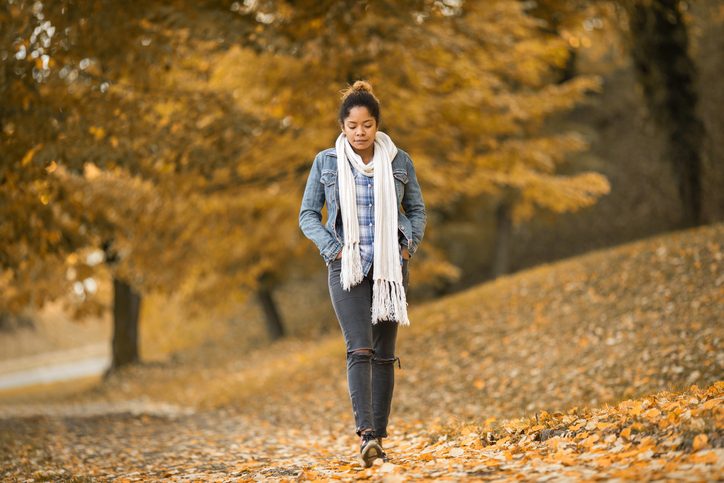 Thoughtful young adult with hair in ponytail, scarf, hands in pockets, walks through park