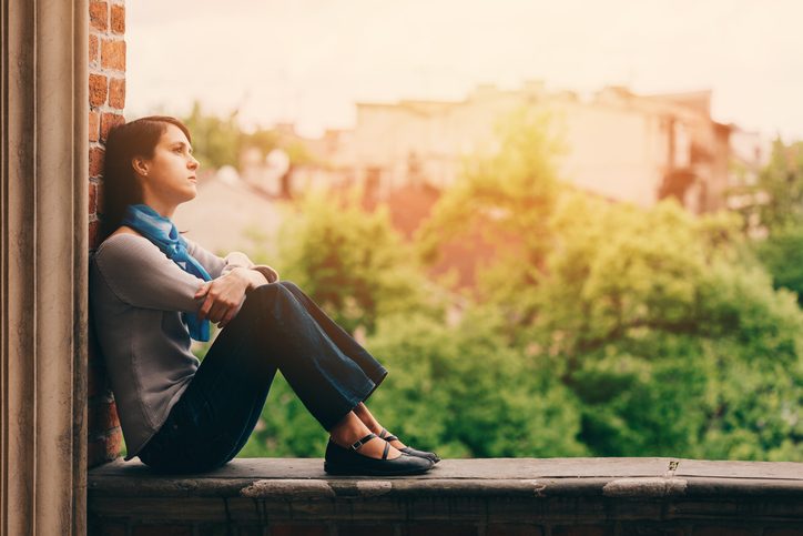 A young person with shoulder-length hair and scarf tied around neck sits on window ledge, looking up, against background of trees and rooftops at sunset