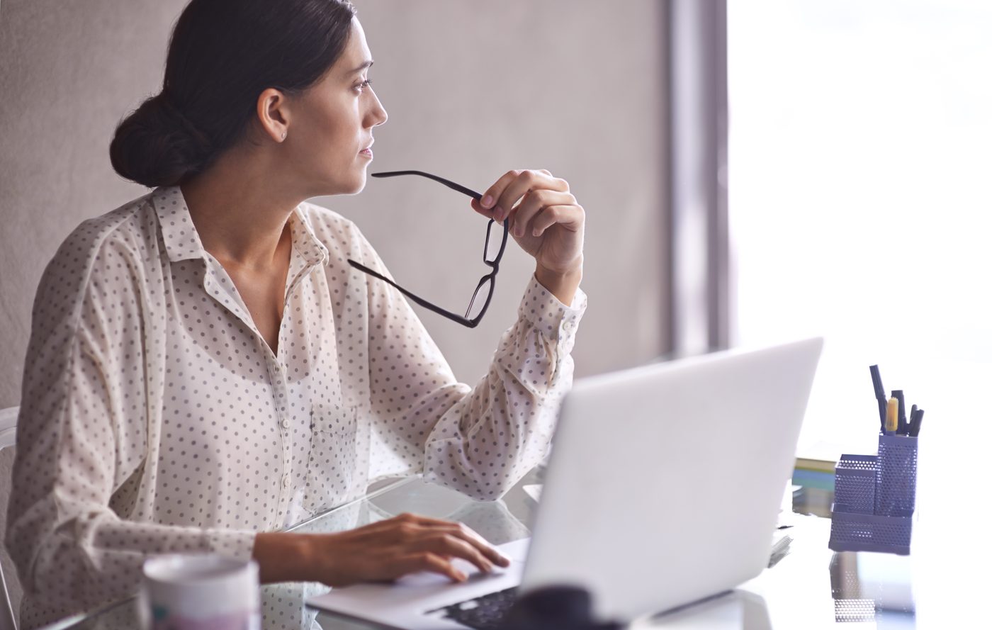 Therapist sits at desk, holding her glasses and looking out the window thoughtfully