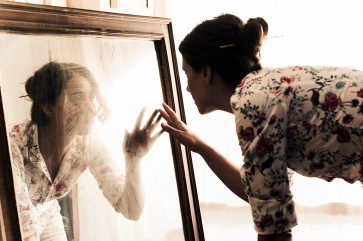 Person leans toward large mirror, fingers to glass. The face is distorted.