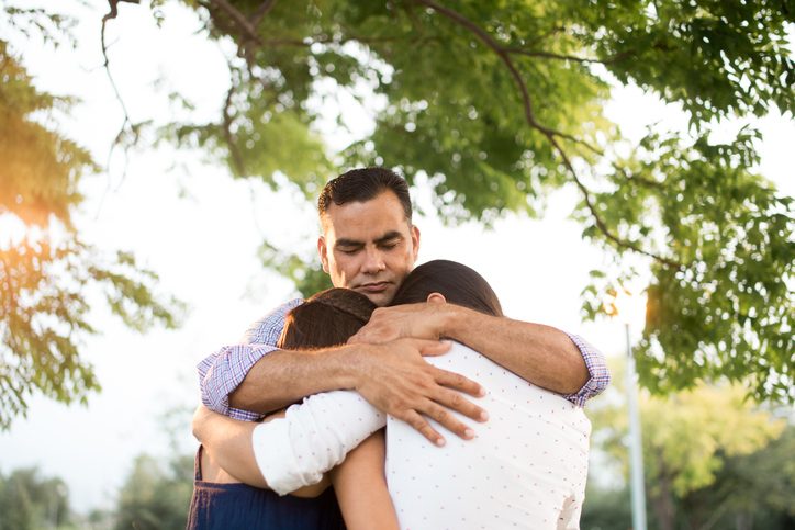 Hispanic parent hugs two teenage children outside under trees