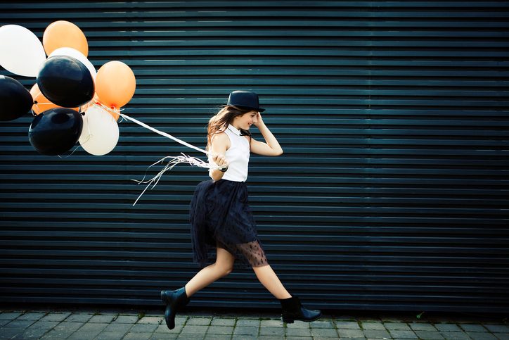 Person with long blonde hair wearing white top, black skirt, and black hat skips along street carrying bunch of Halloween-colored balloons