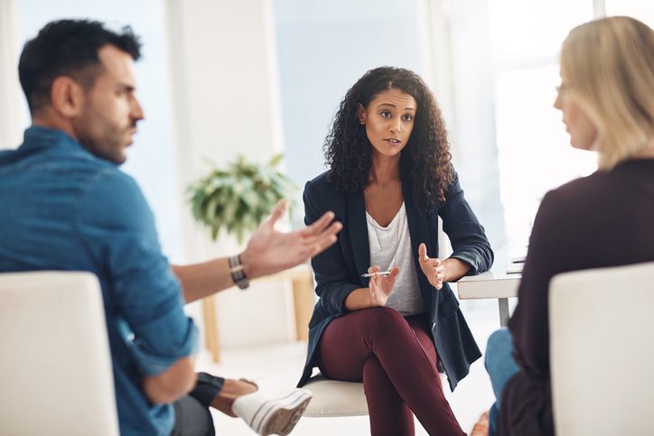 Counselor working with a couple raises hands in gesture while couple talking animatedly featured in rear view