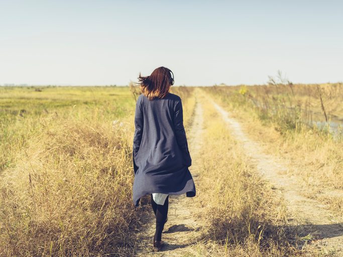 Rear view photo of person with long hair wearing long sweater and headphones walking along path in yellow grassy field