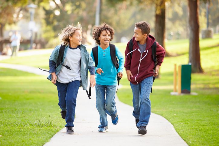 Group of three preteen age kids wearing jeans and backpacks talking happily while walking down path in park