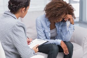 Woman with curly hair sitting on therapist's couch with head leaned on hand. Sitting next to her, a female therapist in a grey coat is listening and taking notes on a clipboard.