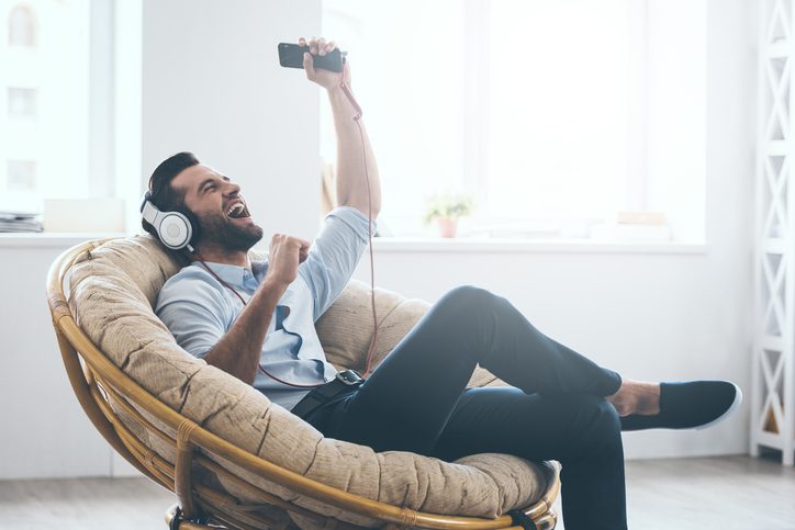 young man in headphones gesturing and keeping eyes closed while listening to music in big comfortable chair at home