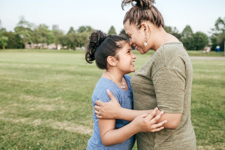 Mother hugs young teen as both laugh together while standing in field outside
