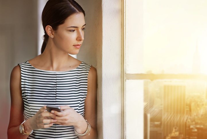 Person in business attire with hair tied back into ponytail holds phone and looks thoughtfully out window