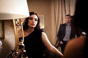 Unhappy looking woman wearing black dress looks in mirror while a man stands in the background