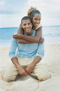 Young black couple on sandy beach. Both are smiling and kneeling on the sand. She has her arms wrapped around his shoulders from behind him.