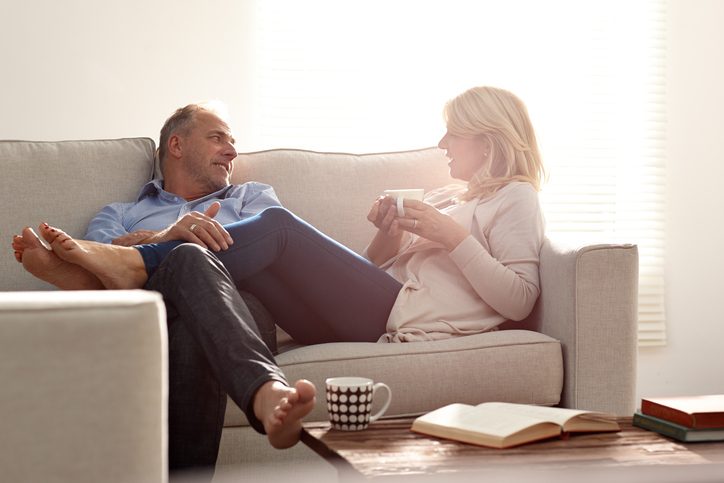 Mature couple relaxing together on sofa hold coffee and smile at each other in living room
