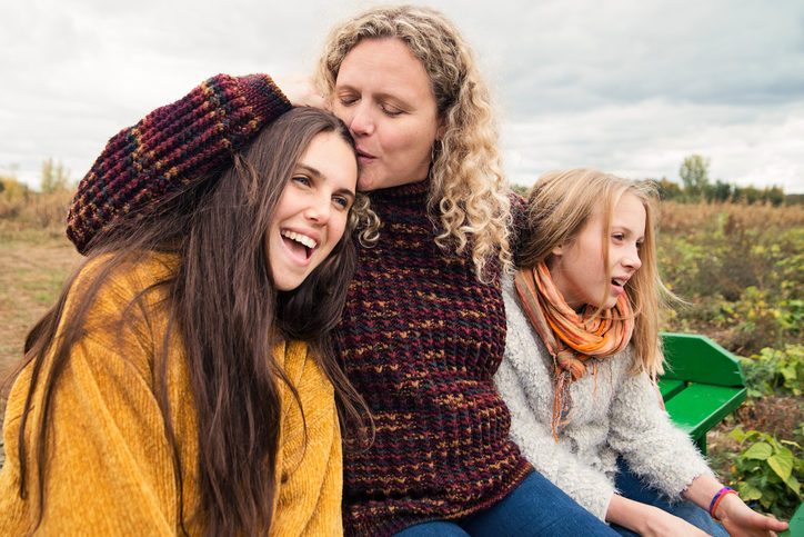 Adult with long curly hair and two adolescents with long hair sit on bench in field looking into distance. Smiling and happy