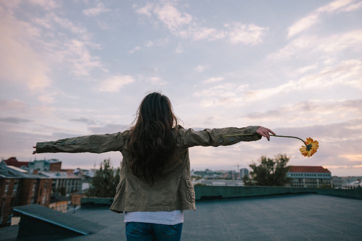 Person stands back on pavement, holding arms out, flower in one hand