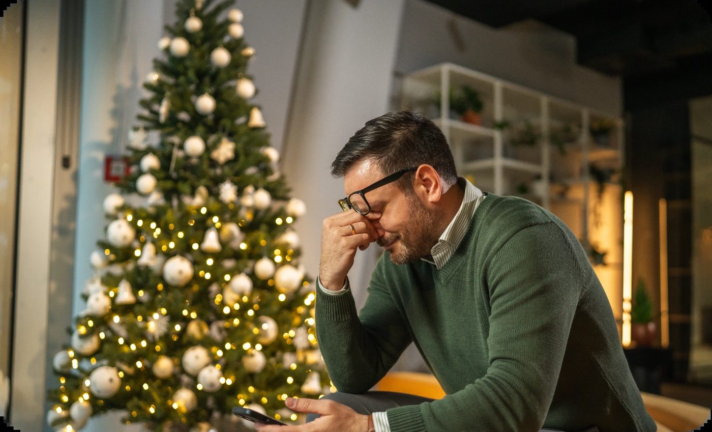 A man with glasses appears stressed or sad, holding his phone near a festive Christmas tree, possibly affected by seasonal affective disorder.