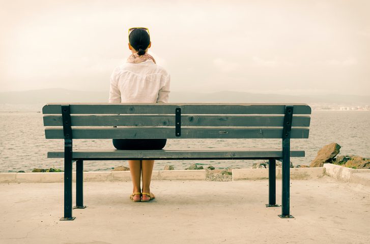 Rear view of a person in white coat, hair in ponytail, sitting on green bench on beach looking out toward the water