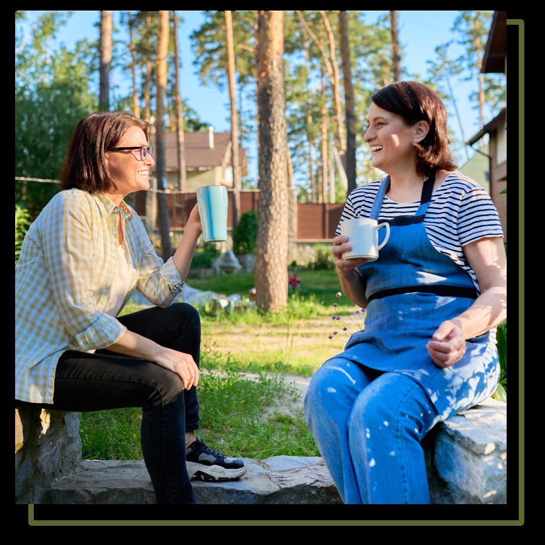 Two middle aged women sitting outdoors together in garden, backyard with mugs of tea