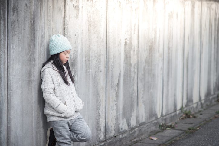 Preteen with long hair under light blue hat stands leaning back against fence, looking thoughtful