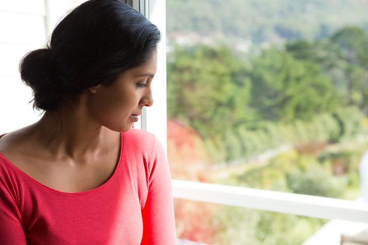 Adult wearing pink top with long hair pulled back looks out window into fields and trees, thoughtful expression on face
