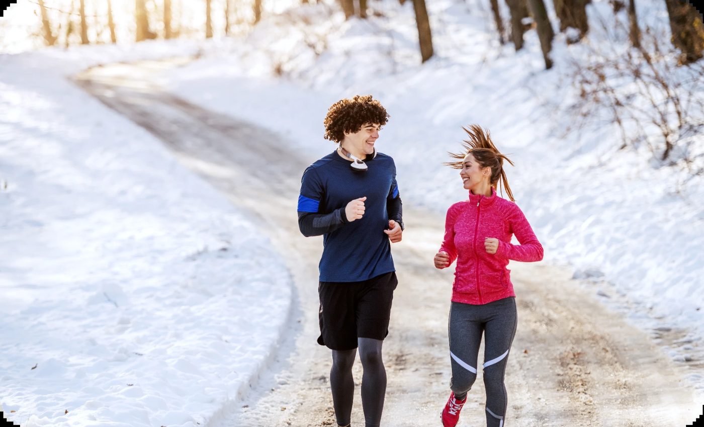 A smiling couple jogs on a snowy path in winter, demonstrating how outdoor exercise can aid in understanding winter depression.
