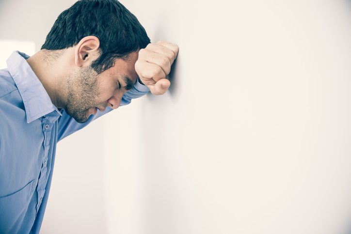 A dark-haired person in button-down shirt leans head against wall